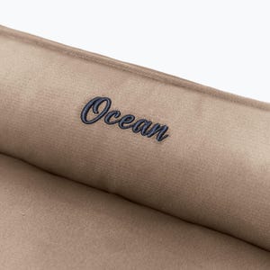 May include: Close-up of a tan fabric with the word "Ocean" embroidered in navy blue script. The fabric appears to be soft and plush, with a subtle sheen. The text is centered on the fabric.