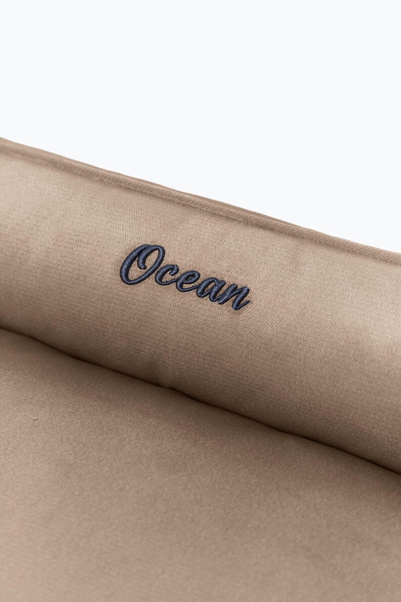 May include: Close-up of a tan fabric with the word "Ocean" embroidered in navy blue script. The fabric appears to be soft and plush, with a subtle sheen. The text is centered on the fabric.