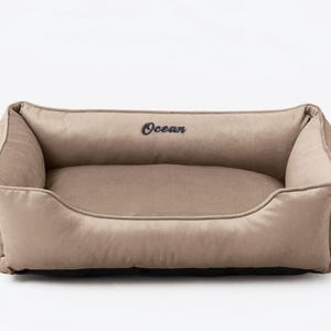 May include: A beige rectangular pet bed with raised sides. The word "Ocean" is embroidered in black on the headrest. The bed has a soft, plush appearance, suggesting comfort for a dog or cat.