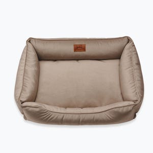 May include: A rectangular dog bed in a neutral beige color. The bed has a plush, cushioned design with raised sides and a lower front. A small, brown leather patch with a logo is sewn on the headrest.
