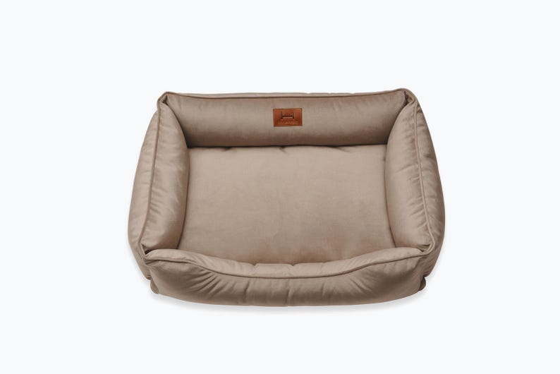 May include: A rectangular dog bed in a neutral beige color. The bed has a plush, cushioned design with raised sides and a lower front. A small, brown leather patch with a logo is sewn on the headrest.