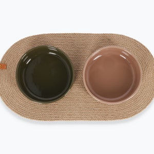 May include: Two ceramic pet bowls, one green and one brown, are placed on a natural fiber oval mat. The mat is made of woven rope and has a leather tag.