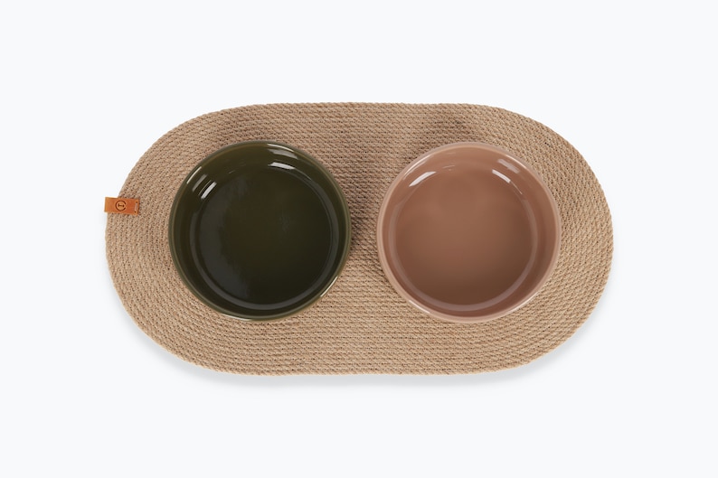 May include: Two ceramic pet bowls, one green and one brown, are placed on a natural fiber oval mat. The mat is made of woven rope and has a leather tag.
