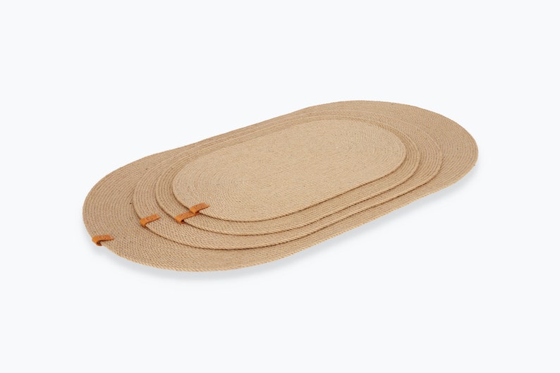 May include: Set of three oval jute placemats with leather trim. The placemats are nested together, with the largest on the bottom and the smallest on top.
