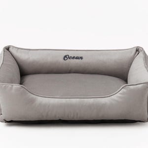 May include: A rectangular, gray pet bed with a plush, velvety texture. The bed features a low front and raised sides for comfort, with the word "Ocean" embroidered in dark blue. The bed is designed for small to medium-sized pets.