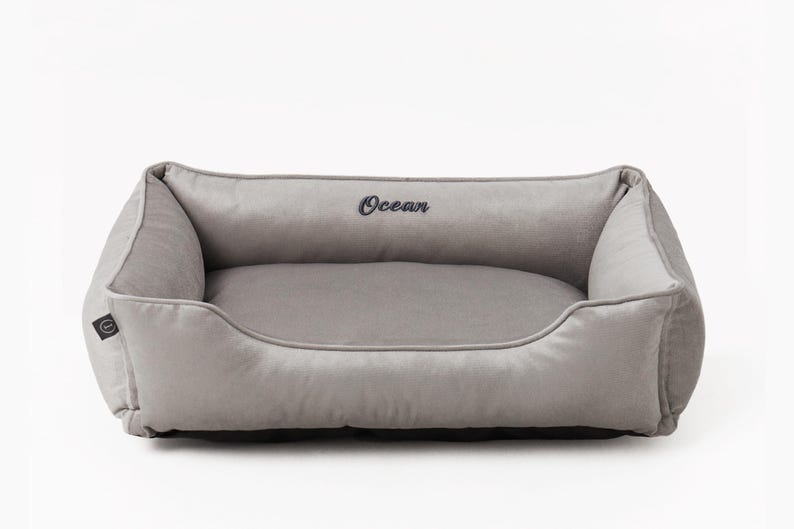 May include: A rectangular, gray pet bed with a plush, velvety texture. The bed features a low front and raised sides for comfort, with the word "Ocean" embroidered in dark blue. The bed is designed for small to medium-sized pets.