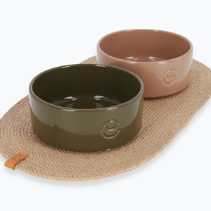 May include: Two ceramic pet bowls, one green and one brown, on a natural fibre mat. The bowls have a raised rim and a small logo on the bottom.