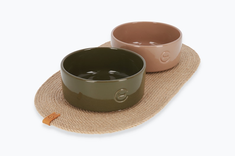 May include: Two ceramic pet bowls, one green and one brown, on a natural fibre mat. The bowls have a raised rim and a small logo on the bottom.