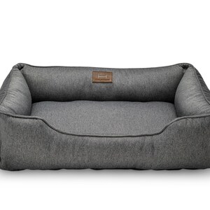 May include: A gray rectangular dog bed with a raised back and sides. The bed has a soft, textured fabric and a brown leather patch with a logo. The bed is designed for comfort and support for pets.