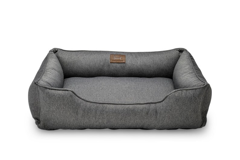 May include: A gray rectangular dog bed with a raised back and sides. The bed has a soft, textured fabric and a brown leather patch with a logo. The bed is designed for comfort and support for pets.