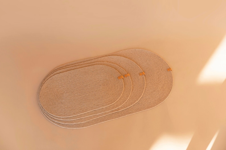 May include: Four beige oval-shaped woven placemats with leather accents. The placemats are stacked on top of each other.