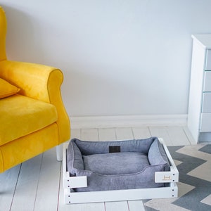May include: A white wooden dog bed with a gray corduroy cushion. The bed is sitting on a white wooden floor with a gray and white chevron rug.