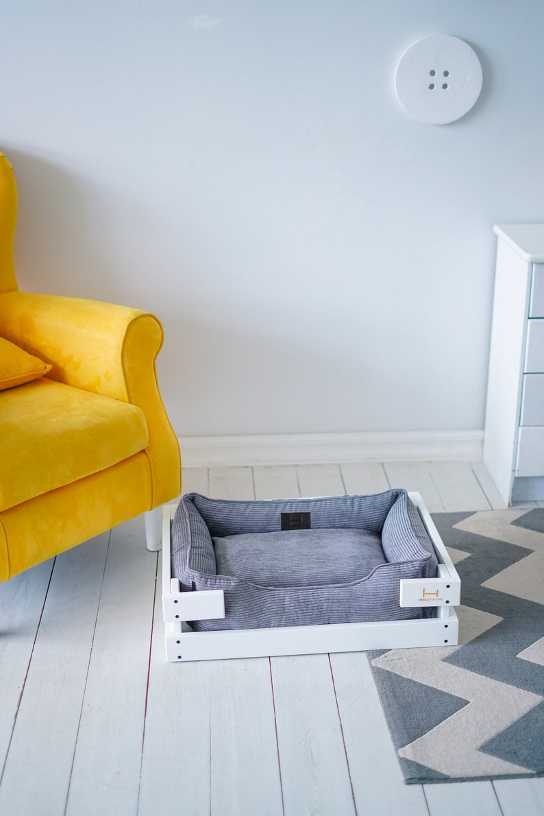 May include: A white wooden dog bed with a gray corduroy cushion. The bed is sitting on a white wooden floor with a gray and white chevron rug.