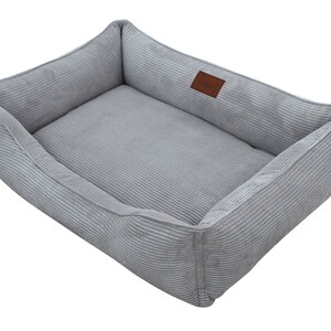 May include: A gray corduroy dog bed with a rectangular shape and a raised edge. The bed has a soft, plush lining and a brown leather label.