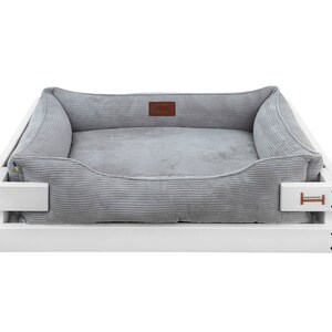 May include: A gray corduroy dog bed with a white wooden frame. The bed has a raised edge and a comfortable, cushioned interior.