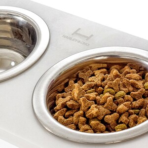 May include: White marble double dog bowl with stainless steel bowls. The bowls are elevated and have a raised rim. One bowl is filled with dry dog food. The marble base has the text "Harley & Cho" engraved on it.