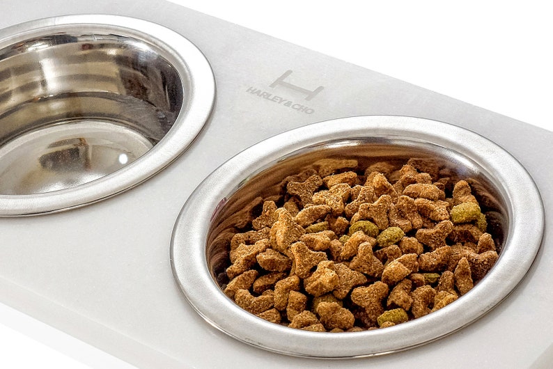 May include: White marble double dog bowl with stainless steel bowls. The bowls are elevated and have a raised rim. One bowl is filled with dry dog food. The marble base has the text "Harley & Cho" engraved on it.