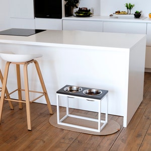 May include: A white elevated dog bowl stand with two stainless steel bowls. The stand is made of metal with a black wood top and is sitting on a light brown wood floor.