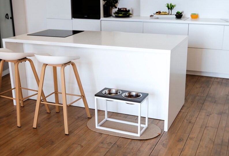 May include: A white elevated dog bowl stand with two stainless steel bowls. The stand is made of metal with a black wood top and is sitting on a light brown wood floor.