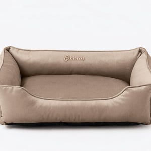 May include: A rectangular dog bed in a neutral beige color. The bed has a plush, cushioned design with raised sides and a low front. The word "Ocean" is embroidered on the back. A small black tag is on the side.