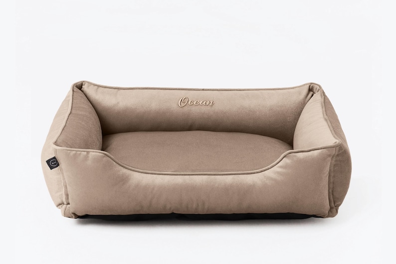 May include: A rectangular dog bed in a neutral beige color. The bed has a plush, cushioned design with raised sides and a low front. The word "Ocean" is embroidered on the back. A small black tag is on the side.
