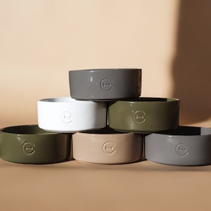 May include: Six ceramic pet bowls stacked in a pyramid shape. The bowls are in various colours, including white, grey, beige, and green. Each bowl has a small, circular logo with the letter "H" in the centre.