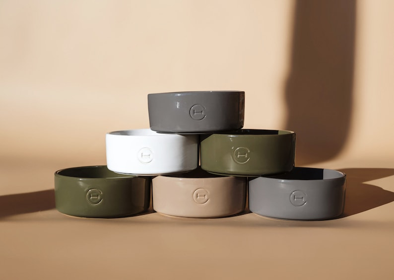May include: Six ceramic pet bowls stacked in a pyramid shape. The bowls are in various colours, including white, grey, beige, and green. Each bowl has a small, circular logo with the letter "H" in the centre.