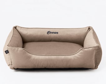 Puede incluir: Una cama beige rectangular para mascotas con laterales elevados. La palabra "Ocean" está bordada en negro en el reposacabezas. La cama tiene un aspecto suave y afelpado, lo que sugiere comodidad para un perro o un gato.
