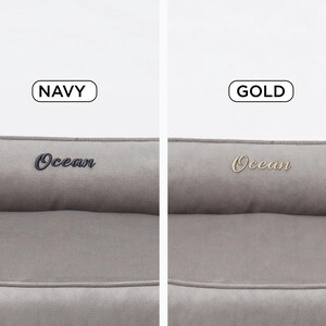 May include: Two pet beds in a neutral gray color. One bed has "Ocean" embroidered in navy blue, and the other has "Ocean" embroidered in gold. The beds have a rectangular shape with a raised bolster.
