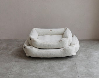 Puede incluir: Una cama para perros blanca y esponjosa con un borde elevado y una cama ligeramente más pequeña en el interior. La cama está hecha de un material suave y afelpado y es perfecta para un perro pequeño.
