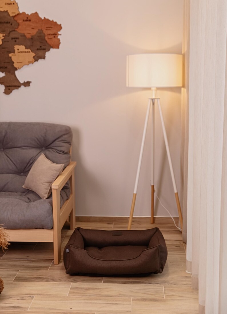May include: A brown dog bed sits on a wooden floor in a living room setting. The bed is rectangular with raised sides. A gray futon, a white floor lamp, and a wooden map of a country are also visible.