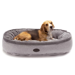 May include: A gray and white oval dog bed with a beagle dog lying in it. The dog bed has a raised edge and a textured fabric.