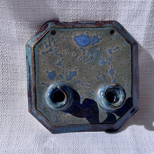 May include: A handmade ceramic wall sconce with a unique octagonal shape. The sconce features a mottled blue and gray glaze with two circular openings for candles. The edges are a darker brown, and it has two holes for hanging.