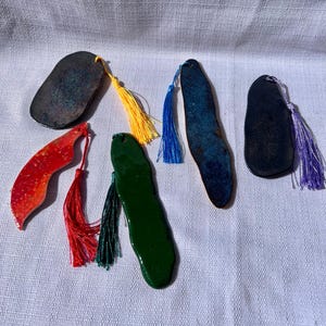 May include: A collection of handmade ceramic bookmarks in various shapes and colors. The bookmarks include a red leaf shape, a green oblong shape, and several dark blue and black oval shapes. Each bookmark has a colorful tassel attached.