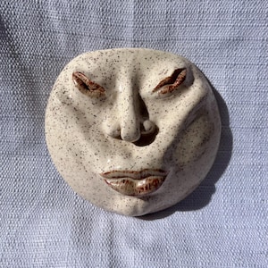 May include: A ceramic face sculpture with closed eyes and a serene expression. The off-white piece has dark speckles and reddish-brown details on the eyes and lips. The round sculpture is set against a light blue textured background.