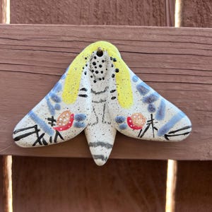 May include: A ceramic moth ornament with a speckled white base, yellow head, and blue wing accents. The wings feature black lines and red floral details. The moth is designed to hang and is suitable for indoor or outdoor decor.