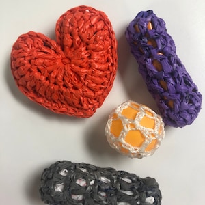 May include: Four crocheted items made from plastic bags. A heart-shaped orange crocheted item, a purple crocheted tube, a yellow crocheted ball, and a grey crocheted tube.