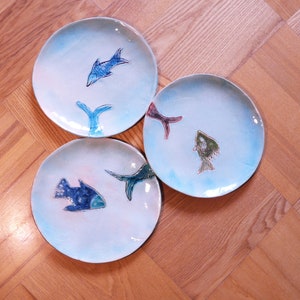 May include: Three white ceramic plates with blue, green, and red fish designs. The plates are stacked on top of each other.