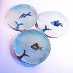 May include: Three ceramic plates with a blue and white glaze. Each plate features a different stylized fish design in shades of blue, green, and brown.