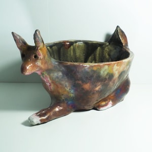 May include: A ceramic bowl shaped like a stylised animal with brown, green, and purple glaze. The bowl has a wide, rounded opening and a narrow base.