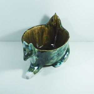 Può includere: Una ciotola in ceramica a forma di cane con smalto verde, marrone e bianco. La ciotola ha un motivo a foglie sul retro.