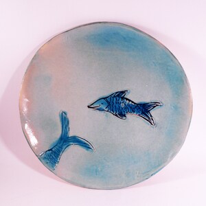 May include: A round ceramic plate with a light blue glaze. The plate features two blue fish, one with its tail visible and the other with its whole body visible. The fish are painted in a simple, stylized manner.