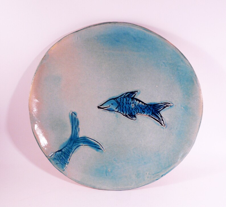 May include: A round ceramic plate with a light blue glaze. The plate features two blue fish, one with its tail visible and the other with its whole body visible. The fish are painted in a simple, stylized manner.