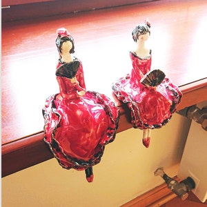 May include: Two ceramic figurines of women in red dresses with black accents and fans. The figurines are posed in a dancing position.