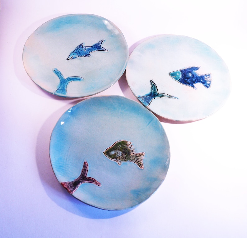 May include: Three ceramic plates with a blue and white glaze. Each plate features a different stylized fish design in shades of blue, green, and brown.