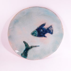May include: A ceramic plate with a blue and green fish design. The fish is swimming towards the right side of the plate. The plate has a light blue background with a pink tint.