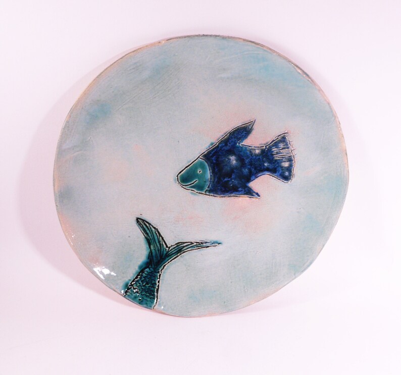 May include: A ceramic plate with a blue and green fish design. The fish is swimming towards the right side of the plate. The plate has a light blue background with a pink tint.