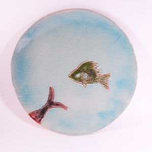 May include: A light blue ceramic plate with a hand-painted design of two fish. One fish is green and brown with a red tail, and the other fish is red and black with a green tail.