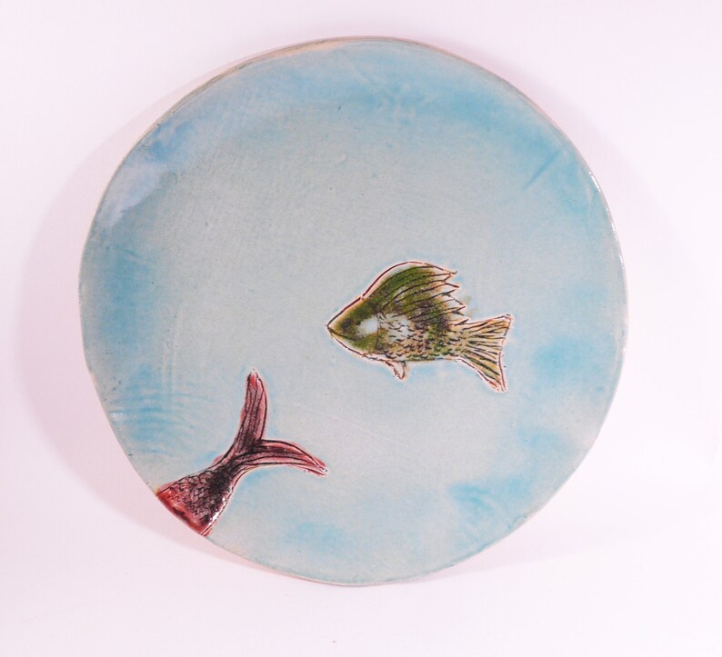 May include: A light blue ceramic plate with a hand-painted design of two fish. One fish is green and brown with a red tail, and the other fish is red and black with a green tail.