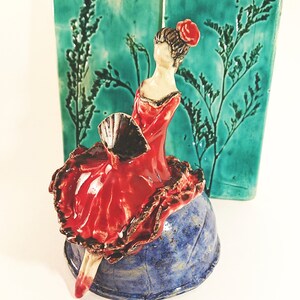 May include: A ceramic figurine of a woman in a red dress and a black fan, sitting on a blue and white ceramic base. The figurine is against a green background with a floral design.